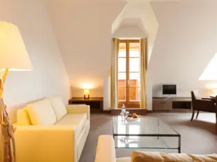 A living area with a light-colored sofa, an armchair and a large glass coffee table. A narrow floor-to-ceiling window, a television and a dark armchair can be seen in the background.