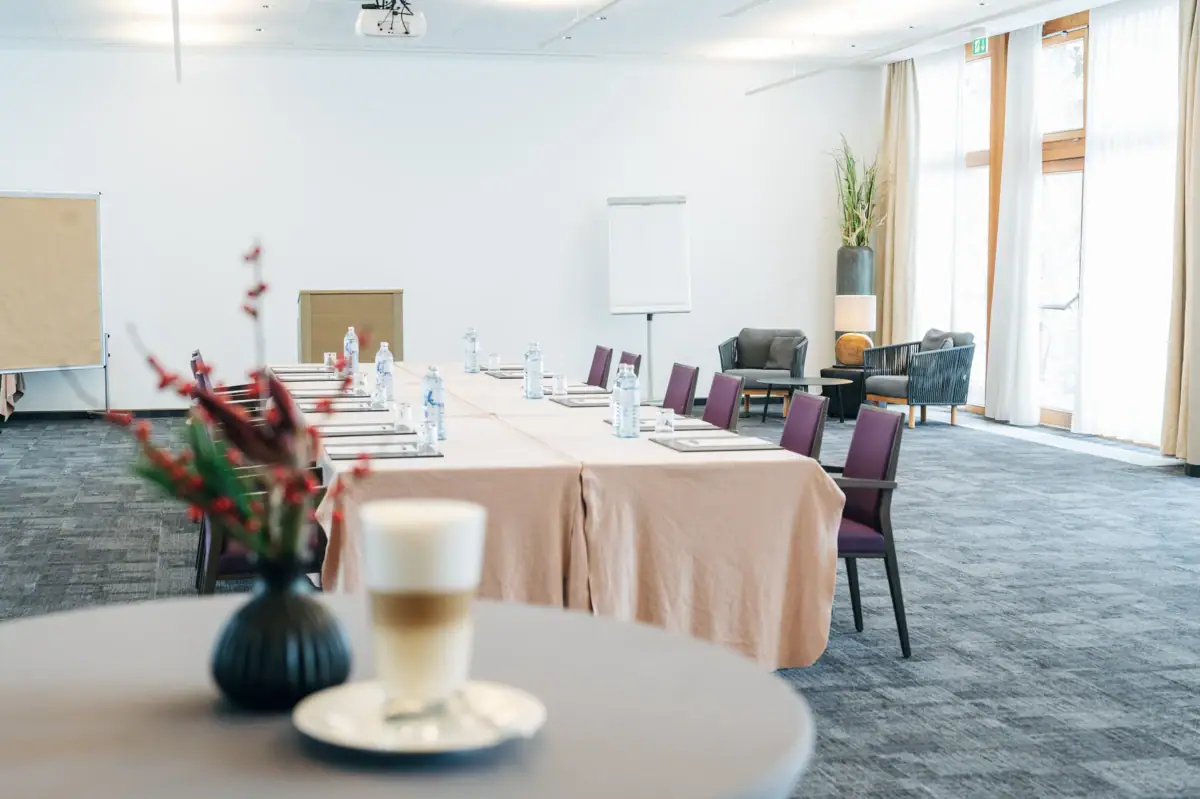 Im Vordergrund ist ein Stehtisch mit einer Blume sowie einem Kaffee zu seheen, während im Hintergrund ein Tagungsraum ist mit einer Blocktafel, um die mehrere Stühle stehen.