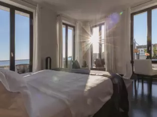 A sun-drenched bedroom with lots of large windows. In the middle of the room is a large, white-covered bed with a view of the sea.  