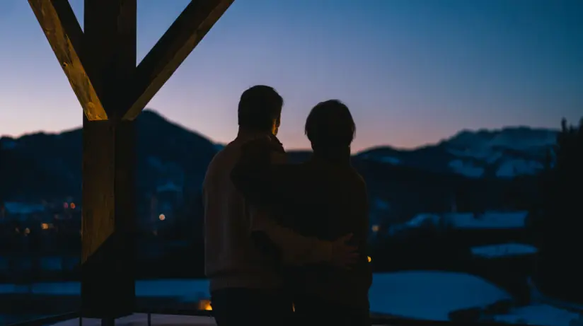 A couple stands on a balcony in the evening darkness and looks into the snow-covered distance