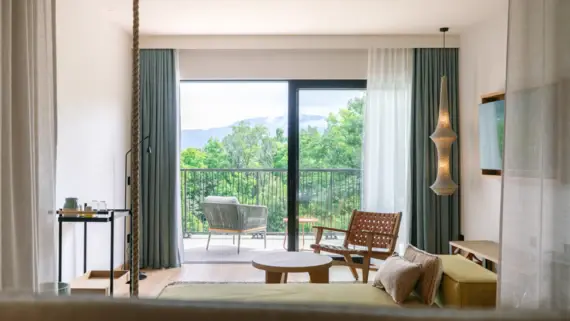 A room with a bed with curtains in the foreground and a chair, as well as another chair on the balcony with a view of the greenery.