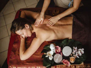 A woman is lying happily and relaxed on a massage table covered by a red cloth and two hands are massaging her back. 