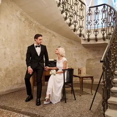 A man in a suit and a woman in a wedding dress sitting on a chair in a room with stairs.