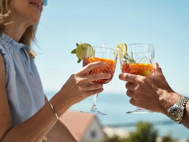 Man and woman outdoors holding drinks in front of the sea in the background.