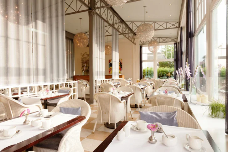 Elegantly set tables with tablecloths, chairs, and vases in a well-lit restaurant interior.