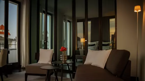 A glass wall separates the front living area with armchairs, side tables and flowers from the rear sleeping area. The colors are kept in a warm grey and light from the setting sun falls into the room through the many large windows.