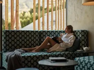 A woman lies on a corner seat while drinking from a cup and holding a book in her hand.