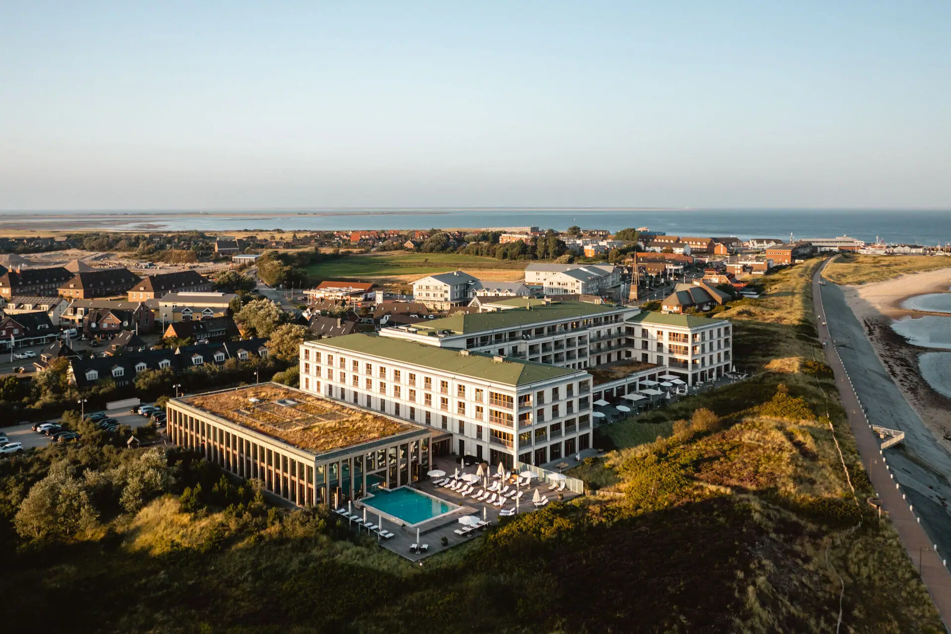 A-ROSA Sylt Eine Luftansicht des A-ROSA Sylt. Das helle Gebäude liegt in der grünen Landschaft und ist nur durch einen Weg vom Meer in der rechten Seite des Bildes getrennt.