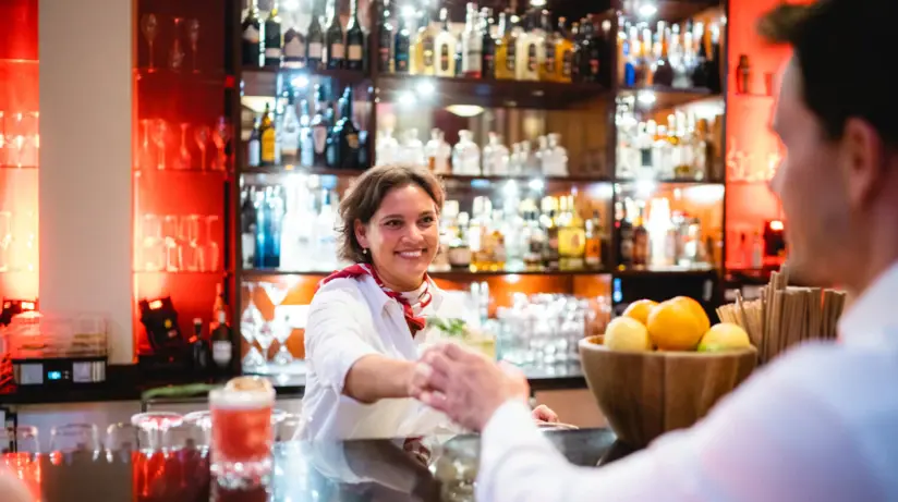 A friendly female bartender hands a cocktail to a customer in a busy bar environment. She wears a white shirt with a red tie and smiles as she slides the drink across the counter. The background of the bar is illuminated with a multitude of bottles of spirits.