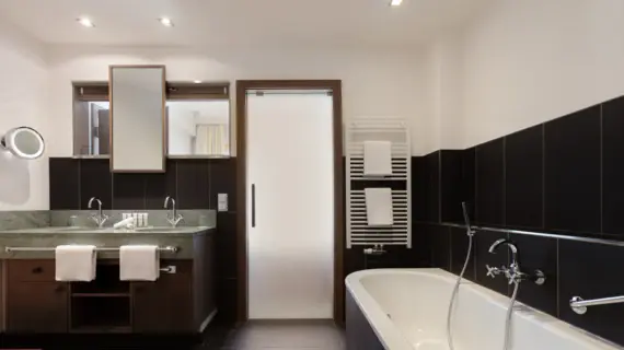 A bathroom with dark tiles, a bathtub and a washstand with two washbasins. A towel warmer with two white towels hangs above the bathtub.