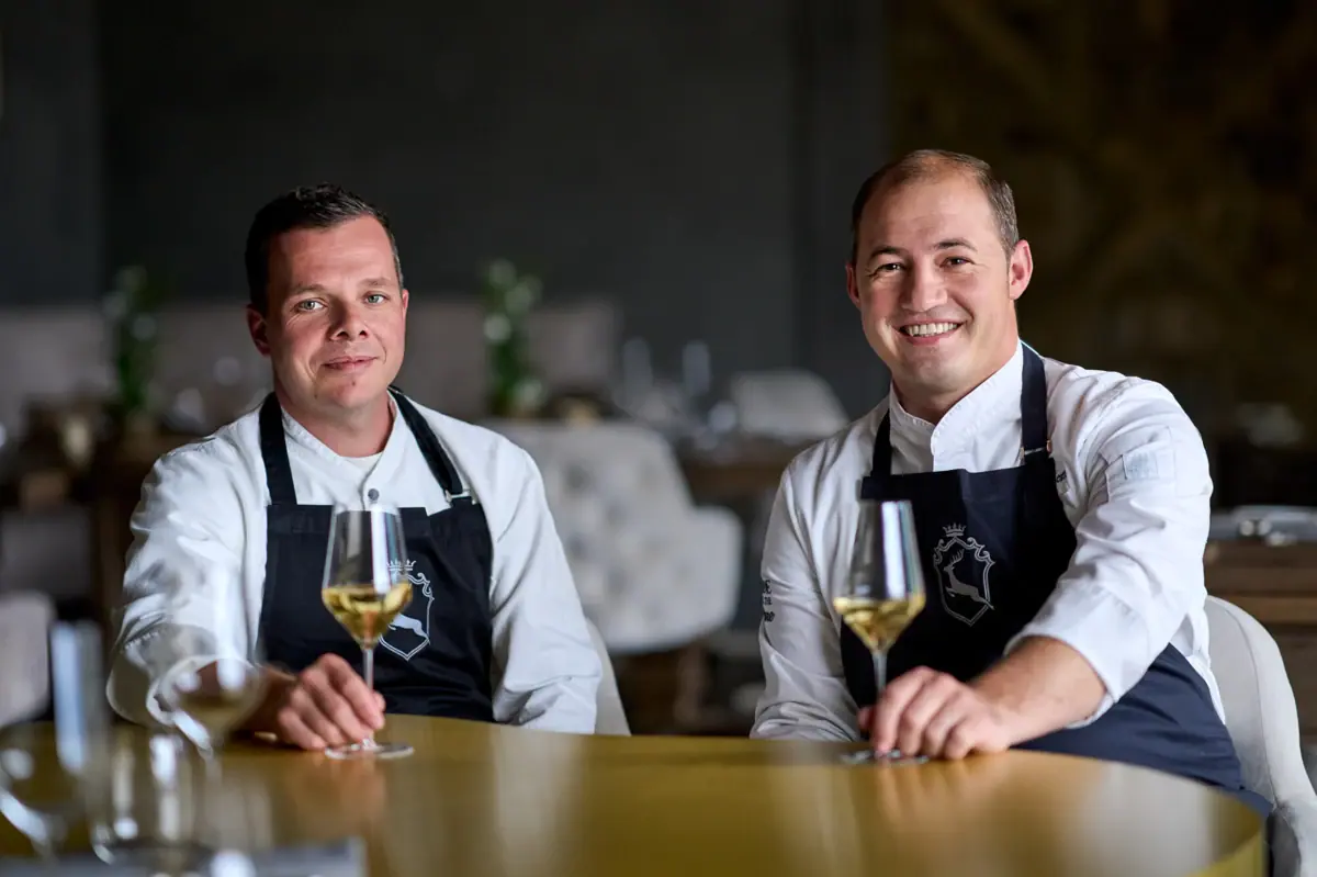 Two men wearing aprons sitting at a table with wine glasses.