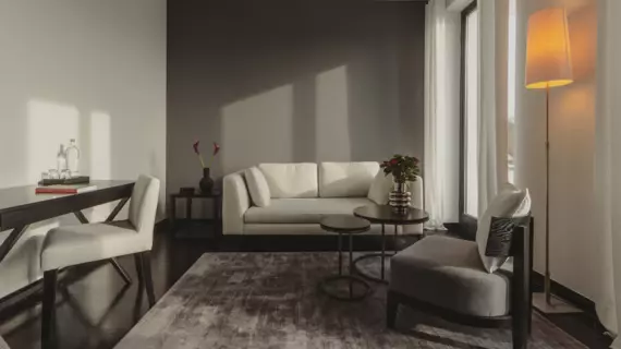 A living area in shades of gray with a light-colored sofa in front of a dark gray wall and side tables with flowers. In front of it is an armchair on a grayish carpet and a lamp emitting warm light. On the left is a desk with a chair.