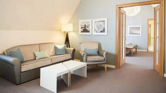 A cozy sitting area in a suite with a sloping roof. A beige couch with patterned cushions and a matching armchair stand next to a simple white coffee table. Two black and white photographs hang on the wall, adding an artistic touch to the room. Through the open wooden door in the background, you can see another part of the room with an upholstered bench and elegant wall decorations that exude an atmosphere of comfort and style.