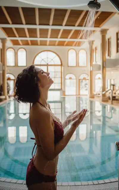 A woman in a wine red bikini is standing under a shower by an indoor pool. 