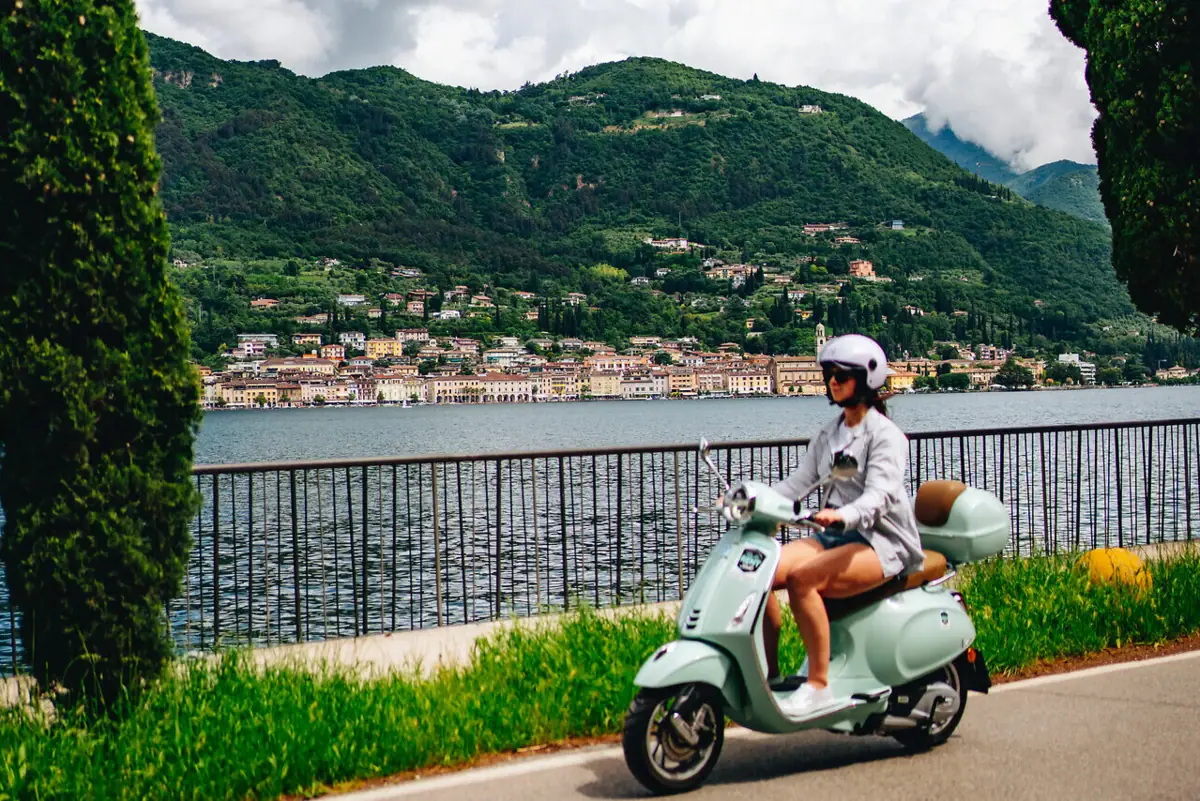 Lake Garda A woman is riding a scooter along a road beside a body of water.