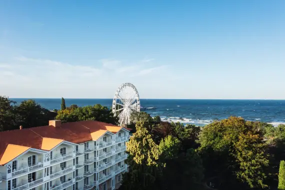 Large hotel building in the style of spa architecture with a red roof between green trees and a white Ferris wheel with the sea in the background.