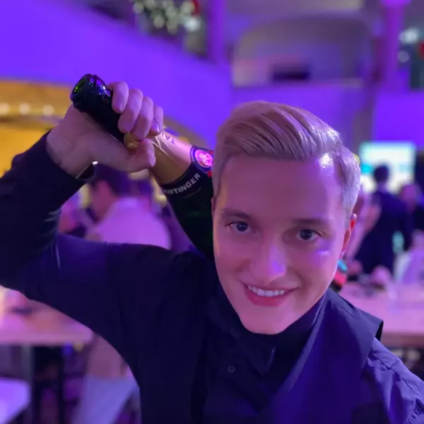 A waiter in uniform smiles at the camera and has a bottle of champagne over his shoulder. 