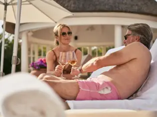 A man and a woman sit on an outdoor lounge lounger with glasses of wine.