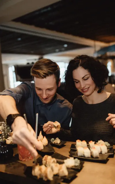 A couple is sitting at a table eating sushi from a big selection of different sushi on dark grey plates.