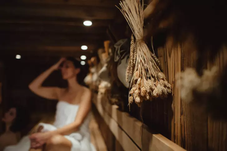 A woman is sitting in a sauna with dry bundles of herbals hanging from the wall. 
