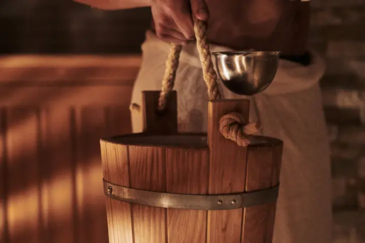 Close-up of a hand holding a wooden sauna bucket, equipped with a metal ladle. The bucket is held by a sturdy rope, which creates an authentic sauna atmosphere. 