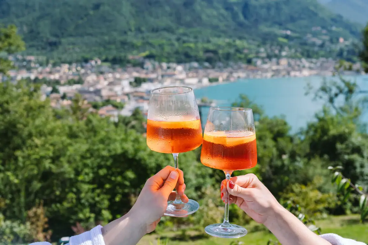 Aperol Spritz Two hands are holding wine glasses filled with an orange-coloured liquid, set against the backdrop of a lake.