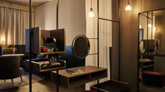 A section of a hotel room with a mirrored wall, in front of which stands a vanity table with a small round mirror. Small lamps hanging from the ceiling fill the room with warm light.