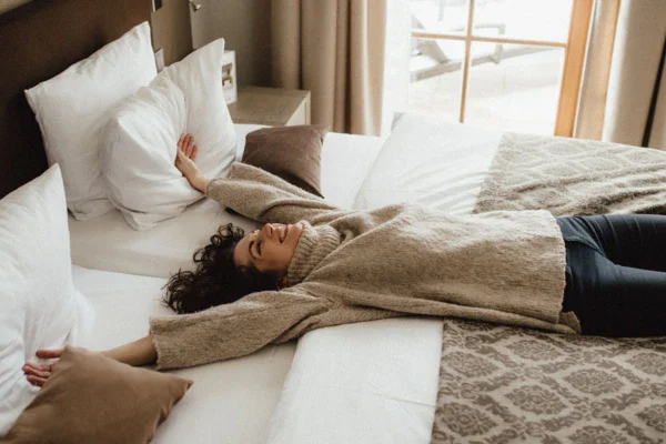 A woman lies happily on a bed in a cozy hotel room and stretches her arms out. 