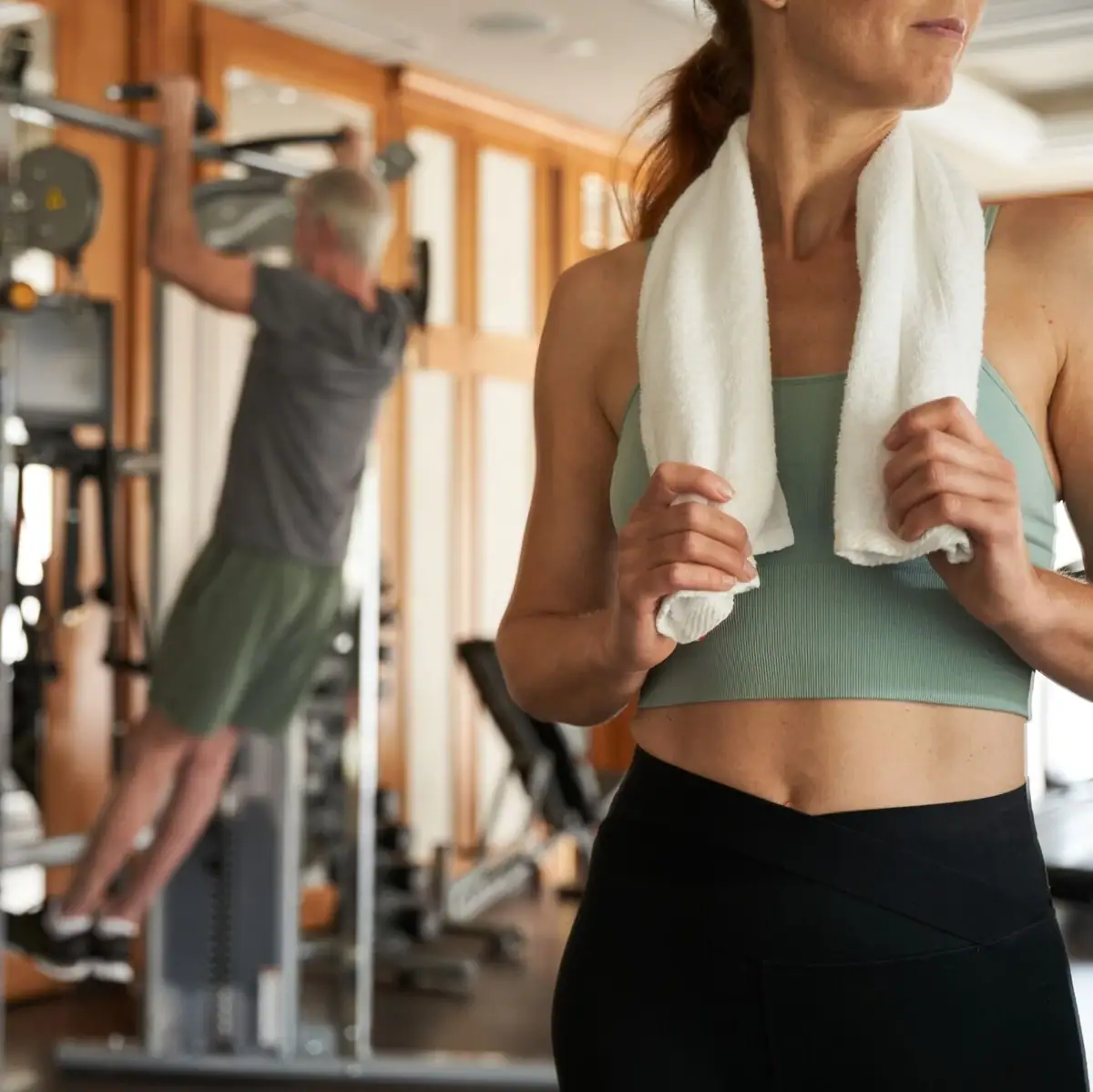 A woman in a gym holding a towel around her neck.