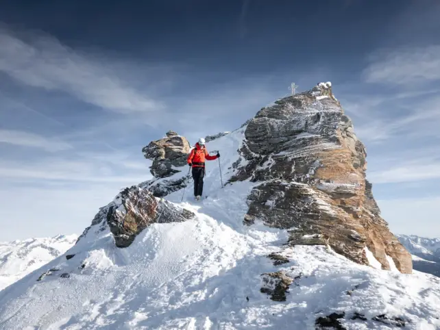 A man in a red jacket and ski equipment walks down a snow-covered peak. 