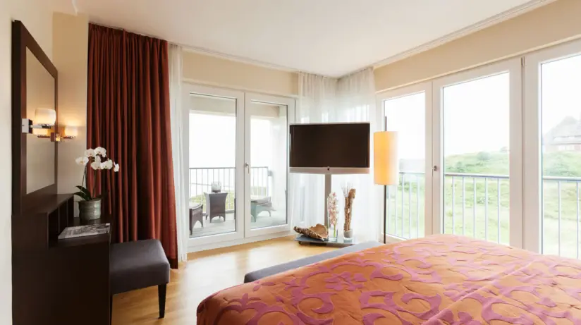 A hotel room with a red and orange bed, a dressing table and television as well as large windows through which the green dunes and the sea can be seen. 