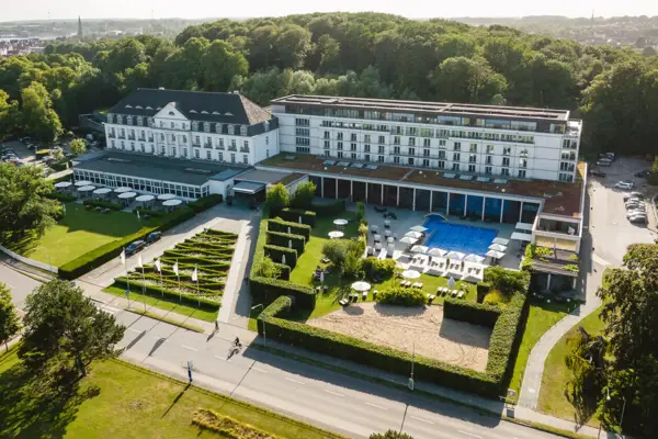Exterior view of the A-ROSA Travemünde with its bright façade and many windows. The harbor and the adjacent sea can be seen in the background. There is a large lawn in front of the building. 