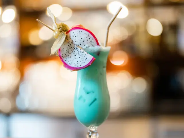 A turquoise cocktail in a tall glass with a slice of dragon fruit and a light straw. 