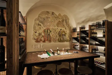 A wine cellar with wine racks on the left and right and a large table in the middle. There are several bottles of wine and wine glasses on the table.