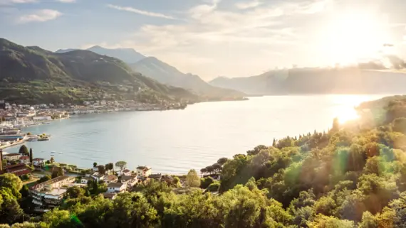 The picture shows an impressive view of Lake Garda, surrounded by a lively town and lush green mountains in the background. The morning sun illuminates the scene, while soft light illuminates the surface of the water and bathes the surrounding landscapes and buildings in a golden glow. This peaceful atmosphere is enhanced by the calm surface of the water and the lush vegetation, which together create an atmosphere of relaxation and closeness to nature.