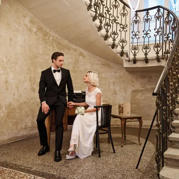 A man in a suit and a woman in a wedding dress sitting on a chair in a room with stairs.