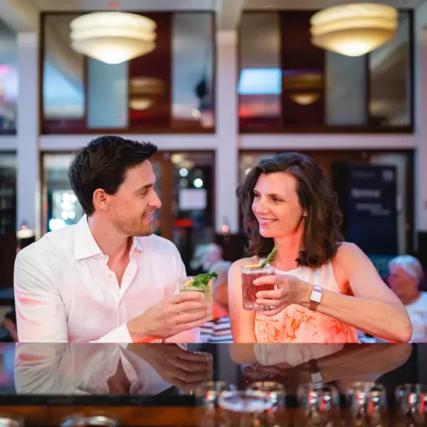 A couple are enjoying a relaxed evening together in a bar. They are sitting at the bar counter, each holding a cocktail in their hand. The man, dressed in a white shirt, smiles at the woman opposite, who is wearing a summery, apricot-colored top and smiles cheerfully at him. 
