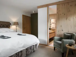 A bedroom with adjoining bathroom. On the left of the picture is the large bed with white bed linen, on which two gray hot water bottles lie and on the right of the picture is a dark stool made of velvet fabric.