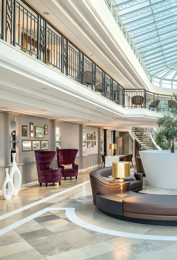 A lobby view of two floors. The upper floor is surrounded by a black and gold fence. A variety of different mirrors hang on the walls and there are various seating options scattered around. 