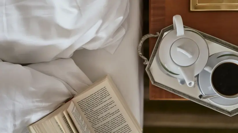 Lamp on a bedside table next to a bed in front of a wall with beige striped wallpaper.