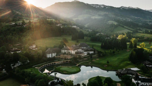 Aerial view of a large building in green surroundings, with a golf course in front and mountains in the background.