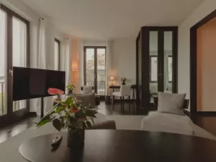 An open-plan living area with large, floor-to-ceiling windows and a comfortable sofa with TV. In the foreground is a table with a vase of flowers. 