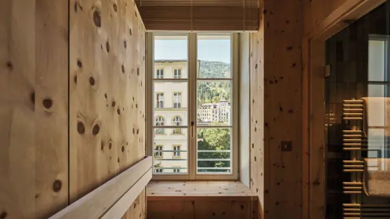 A sauna with a window through which you can see another bright house and a mountain landscape with lots of fir trees.