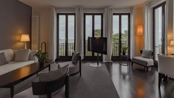 An open-plan living area with floor-to-ceiling windows and light-colored curtains. Small lamps fill the room with sofa, armchairs and TV with warm light. 