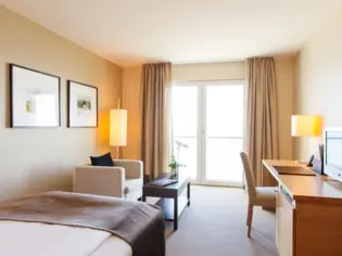 A room with a single bed with a brown bedspread on top. Behind the bed is a light-colored armchair and a coffee table with an orchid on it. A desk and a chair can be seen on the right-hand side of the room.