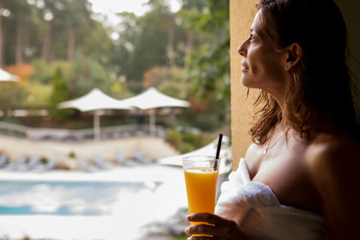 A woman wrapped in a towel sits by the hotel holding a glass with an orange-coloured drink in her hand. 