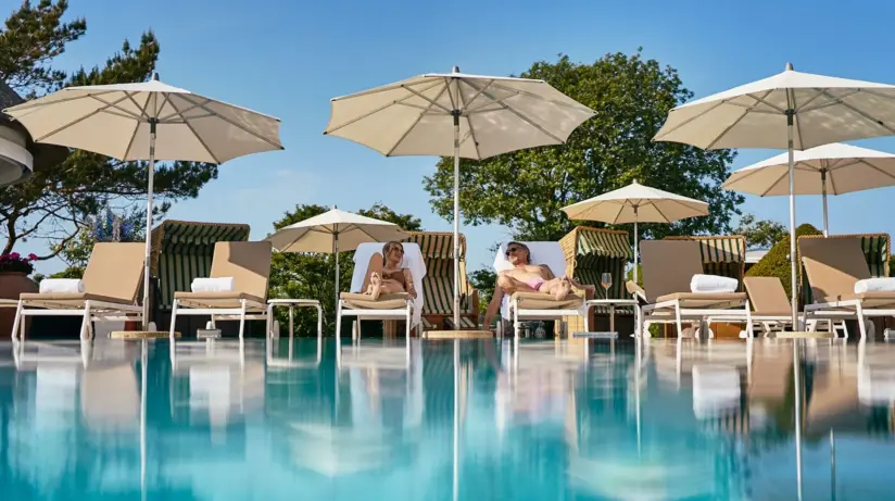 A man and a woman are lying on deckchairs by a pool under a parasol, surrounded by trees.