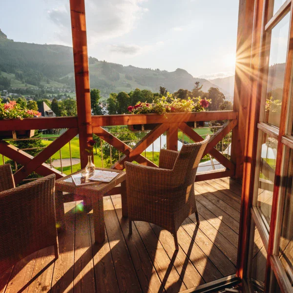 A wooden balcony overlooks a seating area with views of the green landscape and high mountains. The sun rises and shines brightly. 