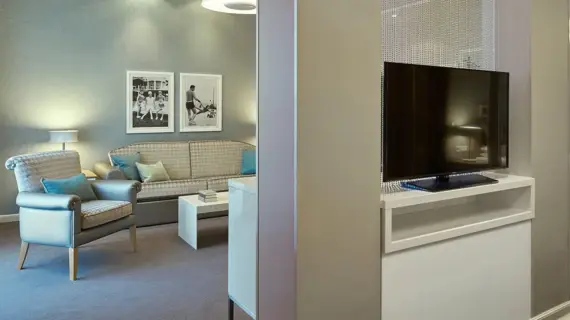 One part of the junior suite features a cozy seating area with a couch and a matching armchair, both in cream and blue tones. Next to the couch is a white coffee table with a few books on it. Two framed black and white photographs hang on the wall above the seating area, exuding elegance and history. On the right of the picture, a modern flat-screen TV can be seen on a simple piece of white furniture, emphasizing the modern comfort of the room.