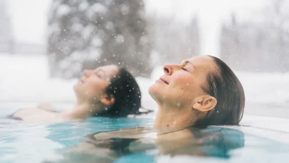 Two women lie back with their eyes closed in a warm, steaming pool and snow falls from the sky.